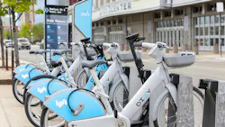 A row of Divvy bike-share bicycles docked at a station at Madison Street and Damen Avenue in Chicago, with the United Center arena visible in the background. The white and light blue bikes display both the Divvy and Lyft logos. A Divvy kiosk with a 'Ride. Return.' information panel stands at the head of the dock. The foreground bikes are electric-assist models with front cargo baskets and integrated locking mechanisms. A row of Divvy bike-share bicycles docked at a station at Madison Street and Damen Avenue in Chicago, with the United Center arena visible in the background. The white and light blue bikes display both the Divvy and Lyft logos. A Divvy kiosk with a 'Ride. Return.' information panel stands at the head of the dock. The foreground bikes are electric-assist models with front cargo baskets and integrated locking mechanisms.