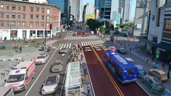 A rendering of a proposed redesign of Flatbush Avenue in Brooklyn, New York, viewed from an elevated angle looking north toward the Manhattan Bridge area. Two dedicated bus-only lanes, marked with dark red pavement and 'BUS ONLY' pavement markings in white, run along the center of the corridor. A blue New York City Transit bus travels in one of the lanes. General traffic lanes flank the bus lanes on both sides, with a police cruiser, ambulance, and several personal vehicles visible. Pedestrians fill the sidewalks and a crosswalk in the foreground. A glass subway entrance structure sits at the median. High-rise buildings and commercial storefronts line both sides of the street under a clear blue sky. A rendering of a proposed redesign of Flatbush Avenue in Brooklyn, New York, viewed from an elevated angle looking north toward the Manhattan Bridge area. Two dedicated bus-only lanes, marked with dark red pavement and 'BUS ONLY' pavement markings in white, run along the center of the corridor. A blue New York City Transit bus travels in one of the lanes. General traffic lanes flank the bus lanes on both sides, with a police cruiser, ambulance, and several personal vehicles visible. Pedestrians fill the sidewalks and a crosswalk in the foreground. A glass subway entrance structure sits at the median. High-rise buildings and commercial storefronts line both sides of the street under a clear blue sky.
