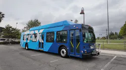 A Fresno Area Express (FAX) transit bus parked in a surface lot, viewed from the front-left at a slight angle. The bus features a two-tone blue livery with large geometric white and light blue graphic elements across the body. A front-mounted bicycle rack is visible. The bus has rooftop-mounted equipment consistent with a compressed natural gas powertrain. A vintage-style lamp post stands nearby, with green athletic fields, trees and an overcast sky in the background.