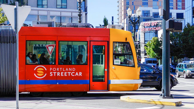 A Portland Streetcar vehicle in its signature red and yellow livery travels through a downtown Portland intersection on a sunny day. The two-tone car displays the Portland Streetcar logo and name on its red mid-section, with a yellow cab at the front marked 'NS' for the North/South line. Several passengers are visible through the windows. A neon 'Go By Streetcar' sign is visible on a building in the background, alongside multi-story mixed-use buildings, ornate street lamps, and light vehicle traffic.