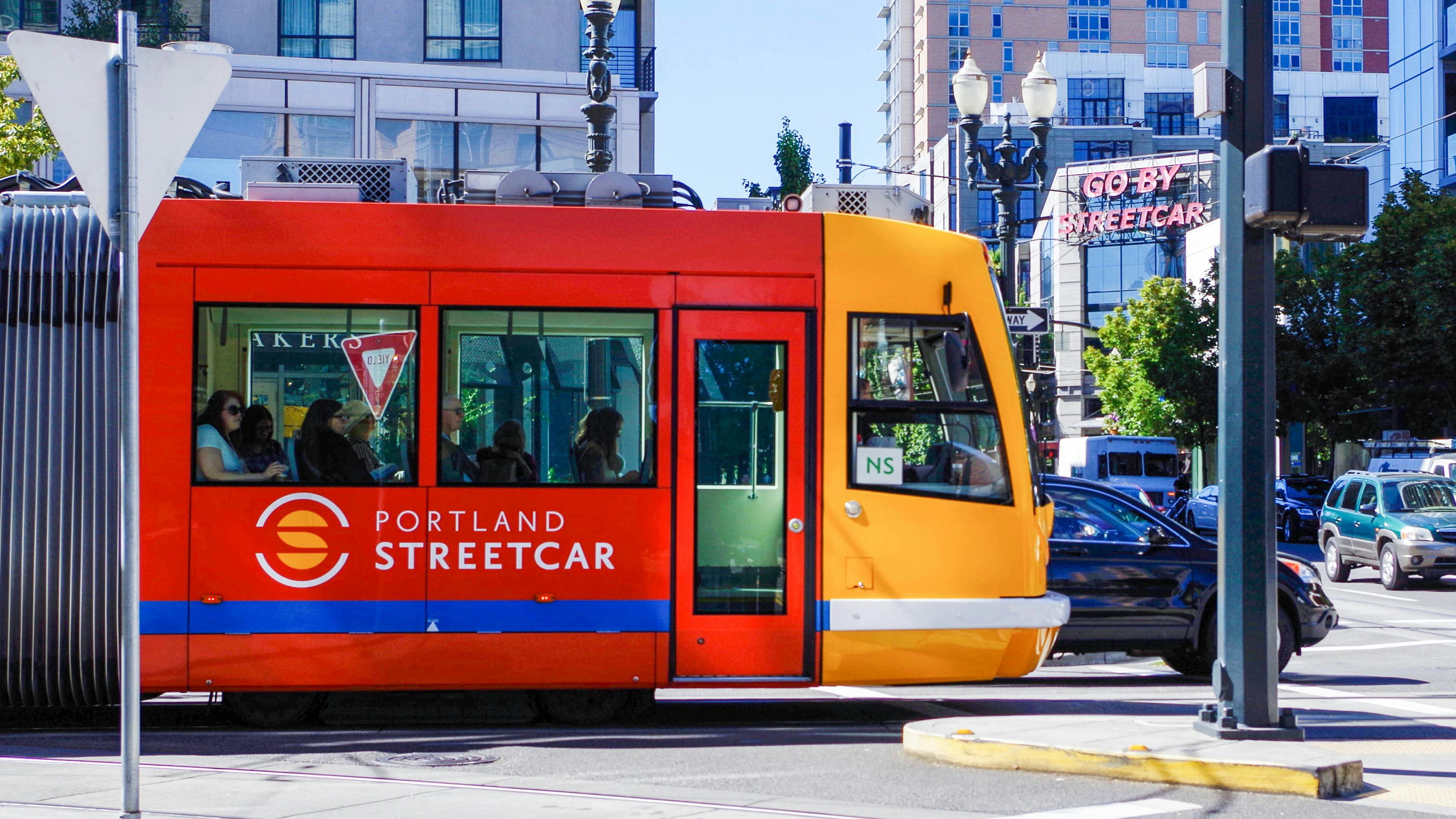 A Portland Streetcar vehicle in its signature red and yellow livery travels through a downtown Portland intersection on a sunny day. The two-tone car displays the Portland Streetcar logo and name on its red mid-section, with a yellow cab at the front marked 'NS' for the North/South line. Several passengers are visible through the windows. A neon 'Go By Streetcar' sign is visible on a building in the background, alongside multi-story mixed-use buildings, ornate street lamps, and light vehicle traffic.