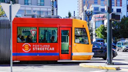 A Portland Streetcar vehicle in its signature red and yellow livery travels through a downtown Portland intersection on a sunny day. The two-tone car displays the Portland Streetcar logo and name on its red mid-section, with a yellow cab at the front marked 'NS' for the North/South line. Several passengers are visible through the windows. A neon 'Go By Streetcar' sign is visible on a building in the background, alongside multi-story mixed-use buildings, ornate street lamps, and light vehicle traffic. A Portland Streetcar vehicle in its signature red and yellow livery travels through a downtown Portland intersection on a sunny day. The two-tone car displays the Portland Streetcar logo and name on its red mid-section, with a yellow cab at the front marked 'NS' for the North/South line. Several passengers are visible through the windows. A neon 'Go By Streetcar' sign is visible on a building in the background, alongside multi-story mixed-use buildings, ornate street lamps, and light vehicle traffic.