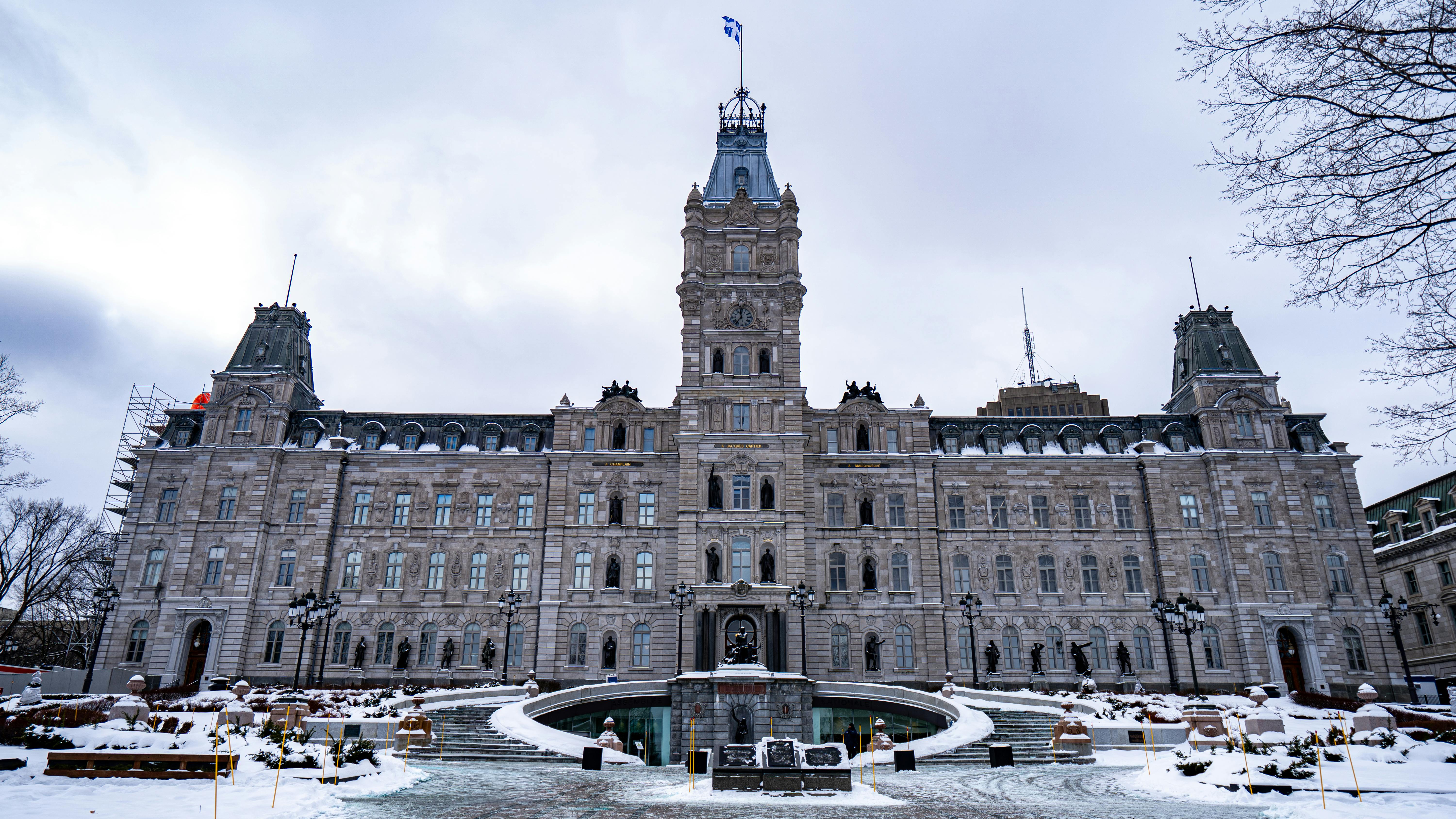 The Quebec National Assembly building photographed from street level on a overcast winter day, with snow covering the grounds and plaza in the foreground. The ornate Second Empire-style stone facade features a central clock tower topped with the Quebec provincial flag, flanked by mansard-roofed corner pavilions. A circular fountain monument anchors the front plaza, flanked by bronze statues of historical figures lining the main approach. Scaffolding is visible on the upper left corner of the building, suggesting active restoration work. Bare trees frame the upper corners of the image against a gray sky.