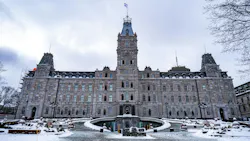 The Quebec National Assembly building photographed from street level on a overcast winter day, with snow covering the grounds and plaza in the foreground. The ornate Second Empire-style stone facade features a central clock tower topped with the Quebec provincial flag, flanked by mansard-roofed corner pavilions. A circular fountain monument anchors the front plaza, flanked by bronze statues of historical figures lining the main approach. Scaffolding is visible on the upper left corner of the building, suggesting active restoration work. Bare trees frame the upper corners of the image against a gray sky. The Quebec National Assembly building photographed from street level on a overcast winter day, with snow covering the grounds and plaza in the foreground. The ornate Second Empire-style stone facade features a central clock tower topped with the Quebec provincial flag, flanked by mansard-roofed corner pavilions. A circular fountain monument anchors the front plaza, flanked by bronze statues of historical figures lining the main approach. Scaffolding is visible on the upper left corner of the building, suggesting active restoration work. Bare trees frame the upper corners of the image against a gray sky.