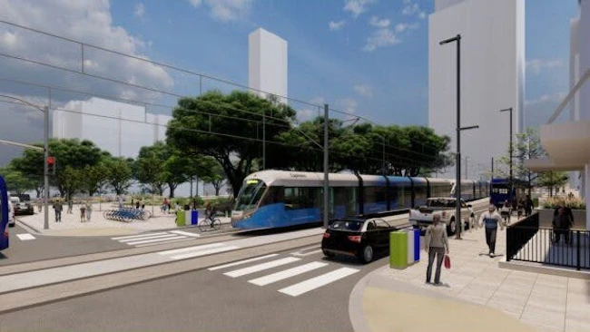 A architectural rendering of a light rail transit station in an urban setting. A blue and silver LRT vehicle travels along a dedicated track through a mixed-use streetscape featuring wide pedestrian plazas, a marked crosswalk in the foreground, mature shade trees, a bike-share station, and modern mid-rise and high-rise buildings in the background under a clear blue sky. Pedestrians and cyclists are visible throughout the scene.