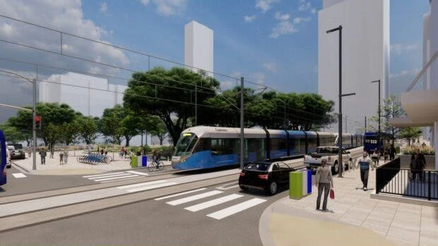 A architectural rendering of a light rail transit station in an urban setting. A blue and silver LRT vehicle travels along a dedicated track through a mixed-use streetscape featuring wide pedestrian plazas, a marked crosswalk in the foreground, mature shade trees, a bike-share station, and modern mid-rise and high-rise buildings in the background under a clear blue sky. Pedestrians and cyclists are visible throughout the scene.