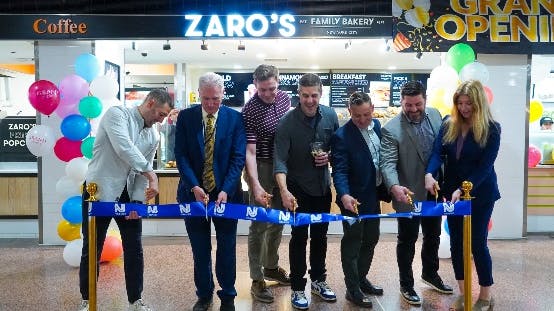 Seven people stand in a line performing a ribbon-cutting ceremony in front of Zaro's Family Bakery, marked by a 'Grand Opening' sign, colorful balloons, and gold stanchions holding a blue ribbon bearing the NJ Transit logo. The group smiles and laughs as they cut the ribbon together inside what appears to be a transit station concourse.