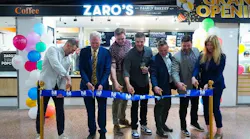 Seven people stand in a line performing a ribbon-cutting ceremony in front of Zaro's Family Bakery, marked by a 'Grand Opening' sign, colorful balloons, and gold stanchions holding a blue ribbon bearing the NJ Transit logo. The group smiles and laughs as they cut the ribbon together inside what appears to be a transit station concourse.