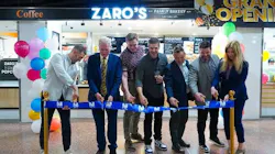 Seven people stand in a line performing a ribbon-cutting ceremony in front of Zaro's Family Bakery, marked by a 'Grand Opening' sign, colorful balloons, and gold stanchions holding a blue ribbon bearing the NJ Transit logo. The group smiles and laughs as they cut the ribbon together inside what appears to be a transit station concourse. Seven people stand in a line performing a ribbon-cutting ceremony in front of Zaro's Family Bakery, marked by a 'Grand Opening' sign, colorful balloons, and gold stanchions holding a blue ribbon bearing the NJ Transit logo. The group smiles and laughs as they cut the ribbon together inside what appears to be a transit station concourse.