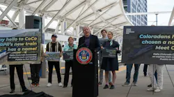 Colorado Governor Jared Polis speaks at a podium bearing the State of Colorado seal at an outdoor transit station platform, announcing the name of the Front Range Passenger Rail's new express train. Supporters standing behind him hold signs reading 'Say hello to CoCo — FRPR's new train has a name!' and 'Coloradans have chosen a name! Meet CoCo,' describing CoCo (Colorado Connector) as an express train. A large canopy structure and high-rise building are visible in the background.