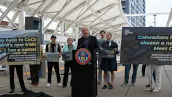 Colorado Governor Jared Polis speaks at a podium bearing the State of Colorado seal at an outdoor transit station platform, announcing the name of the Front Range Passenger Rail's new express train. Supporters standing behind him hold signs reading 'Say hello to CoCo — FRPR's new train has a name!' and 'Coloradans have chosen a name! Meet CoCo,' describing CoCo (Colorado Connector) as an express train. A large canopy structure and high-rise building are visible in the background. Colorado Governor Jared Polis speaks at a podium bearing the State of Colorado seal at an outdoor transit station platform, announcing the name of the Front Range Passenger Rail's new express train. Supporters standing behind him hold signs reading 'Say hello to CoCo — FRPR's new train has a name!' and 'Coloradans have chosen a name! Meet CoCo,' describing CoCo (Colorado Connector) as an express train. A large canopy structure and high-rise building are visible in the background.