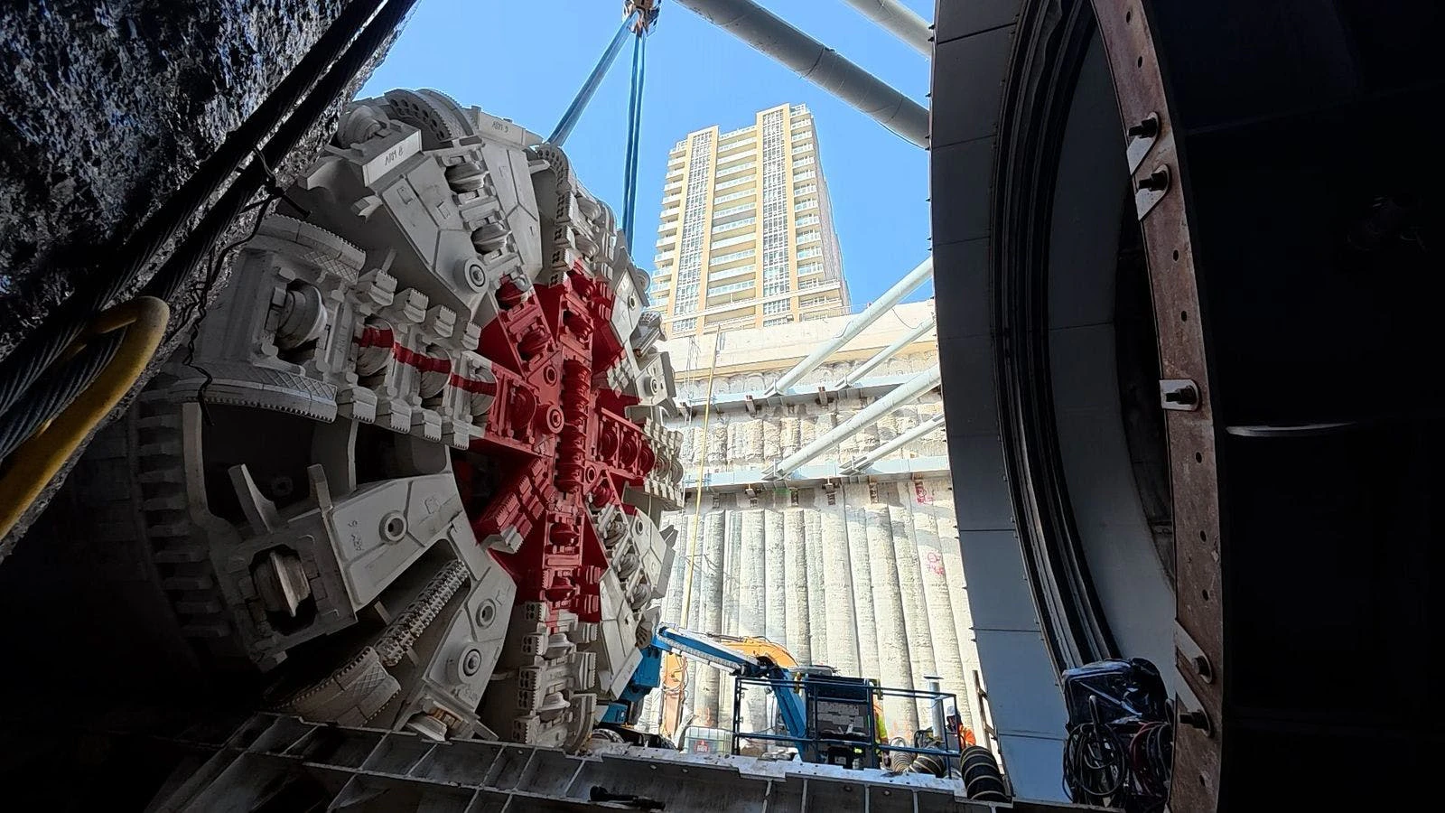 A tunnel boring machine (TBM) cutterhead is lowered by crane into an open construction shaft in an urban setting. The circular cutterhead, featuring a white-and-red face with radial spokes and rows of disc cutters, is suspended mid-air against a backdrop of a concrete retaining wall and a tall residential high-rise tower under a clear blue sky. Construction equipment including a crane and lift platform are visible at the base of the shaft. The circular steel entry portal of the tunnel is visible at right.