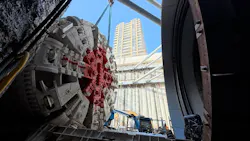 A tunnel boring machine (TBM) cutterhead is lowered by crane into an open construction shaft in an urban setting. The circular cutterhead, featuring a white-and-red face with radial spokes and rows of disc cutters, is suspended mid-air against a backdrop of a concrete retaining wall and a tall residential high-rise tower under a clear blue sky. Construction equipment including a crane and lift platform are visible at the base of the shaft. The circular steel entry portal of the tunnel is visible at right. A tunnel boring machine (TBM) cutterhead is lowered by crane into an open construction shaft in an urban setting. The circular cutterhead, featuring a white-and-red face with radial spokes and rows of disc cutters, is suspended mid-air against a backdrop of a concrete retaining wall and a tall residential high-rise tower under a clear blue sky. Construction equipment including a crane and lift platform are visible at the base of the shaft. The circular steel entry portal of the tunnel is visible at right.