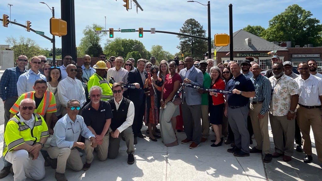 A large group of approximately 40 officials, staff, and construction workers pose together at a ribbon-cutting ceremony at a busy intersection on Columbia Pike in Arlington, Virginia. Several people in the front hold a black ribbon bearing the Arlington County logo. Attendees include people in business attire and construction workers in high-visibility vests and hard hats. A traffic signal arm extends overhead, with a green 'Columbia Pike' street sign visible in the background. Trees, commercial storefronts including a Celtic House business, and a clear blue sky are visible behind the group.