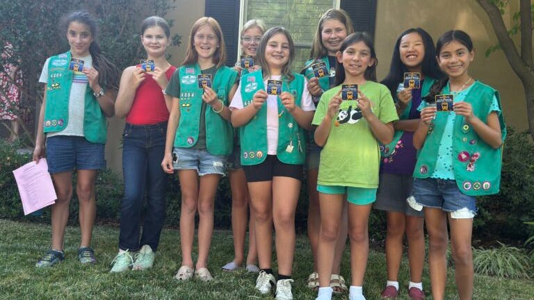 A group of nine children, most wearing green Girl Scout vests adorned with badges, pose outdoors on a grass lawn and hold up transit-related cards or badges earned through a transit agency scout program. The children smile at the camera in front of a residential building with trees and shrubs in the background on a sunny day.
