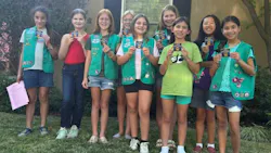 A group of nine children, most wearing green Girl Scout vests adorned with badges, pose outdoors on a grass lawn and hold up transit-related cards or badges earned through a transit agency scout program. The children smile at the camera in front of a residential building with trees and shrubs in the background on a sunny day. A group of nine children, most wearing green Girl Scout vests adorned with badges, pose outdoors on a grass lawn and hold up transit-related cards or badges earned through a transit agency scout program. The children smile at the camera in front of a residential building with trees and shrubs in the background on a sunny day.