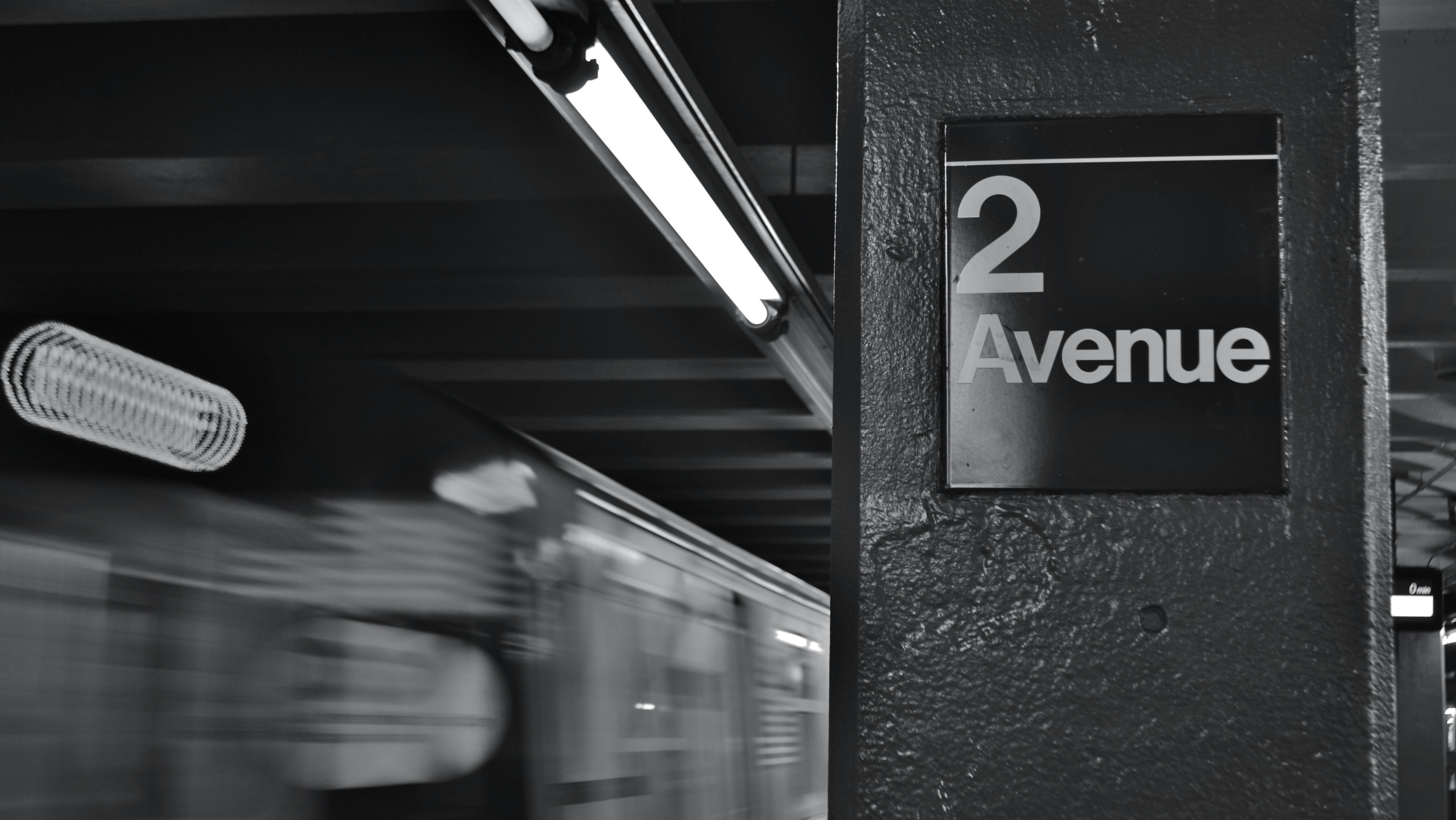A black-and-white photo of a New York City subway platform column bearing a '2 Avenue' station sign, with a motion-blurred train passing in the background. Fluorescent lights run along the low ceiling above the platform.