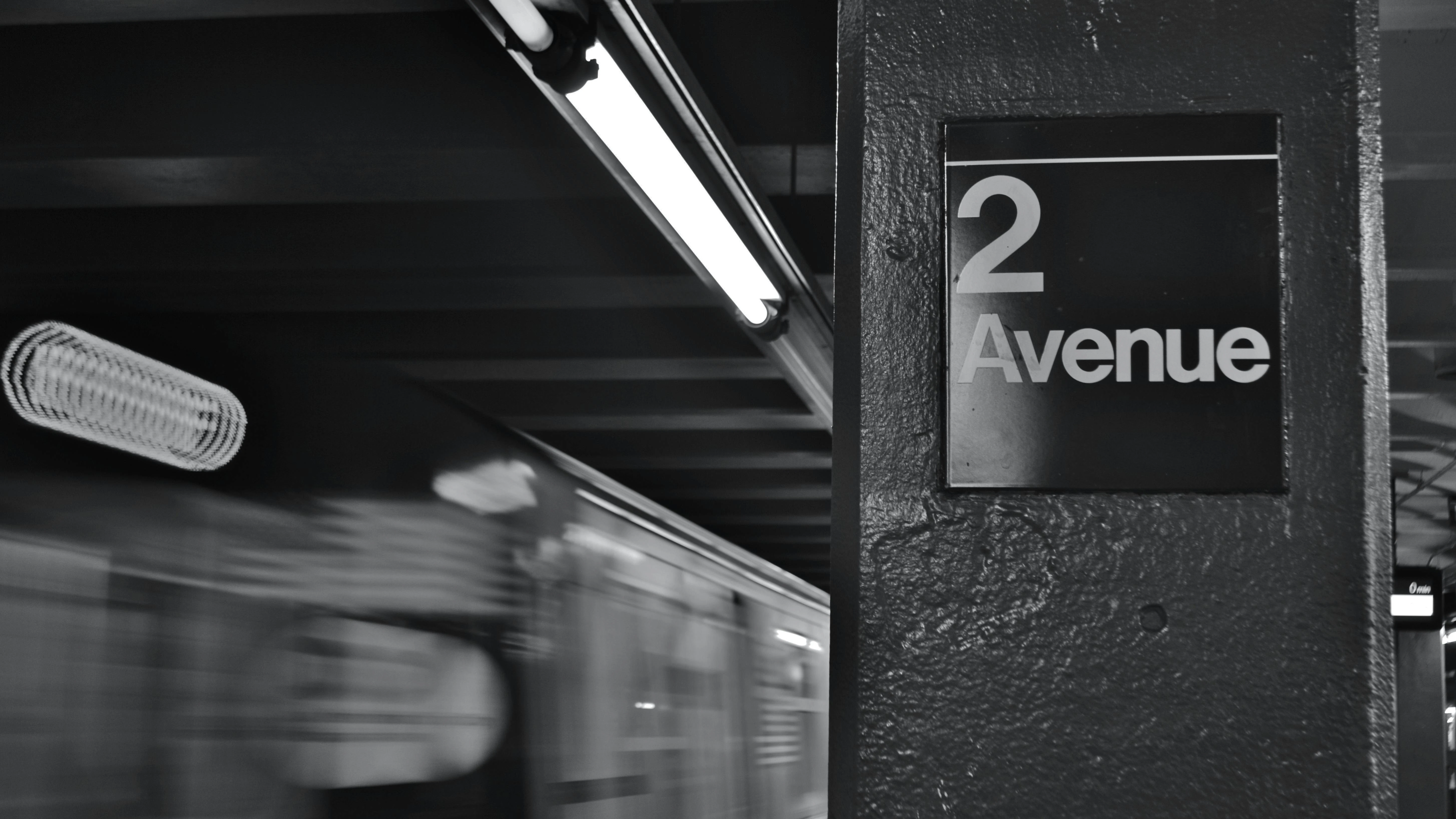 A black-and-white photo of a New York City subway platform column bearing a '2 Avenue' station sign, with a motion-blurred train passing in the background. Fluorescent lights run along the low ceiling above the platform.