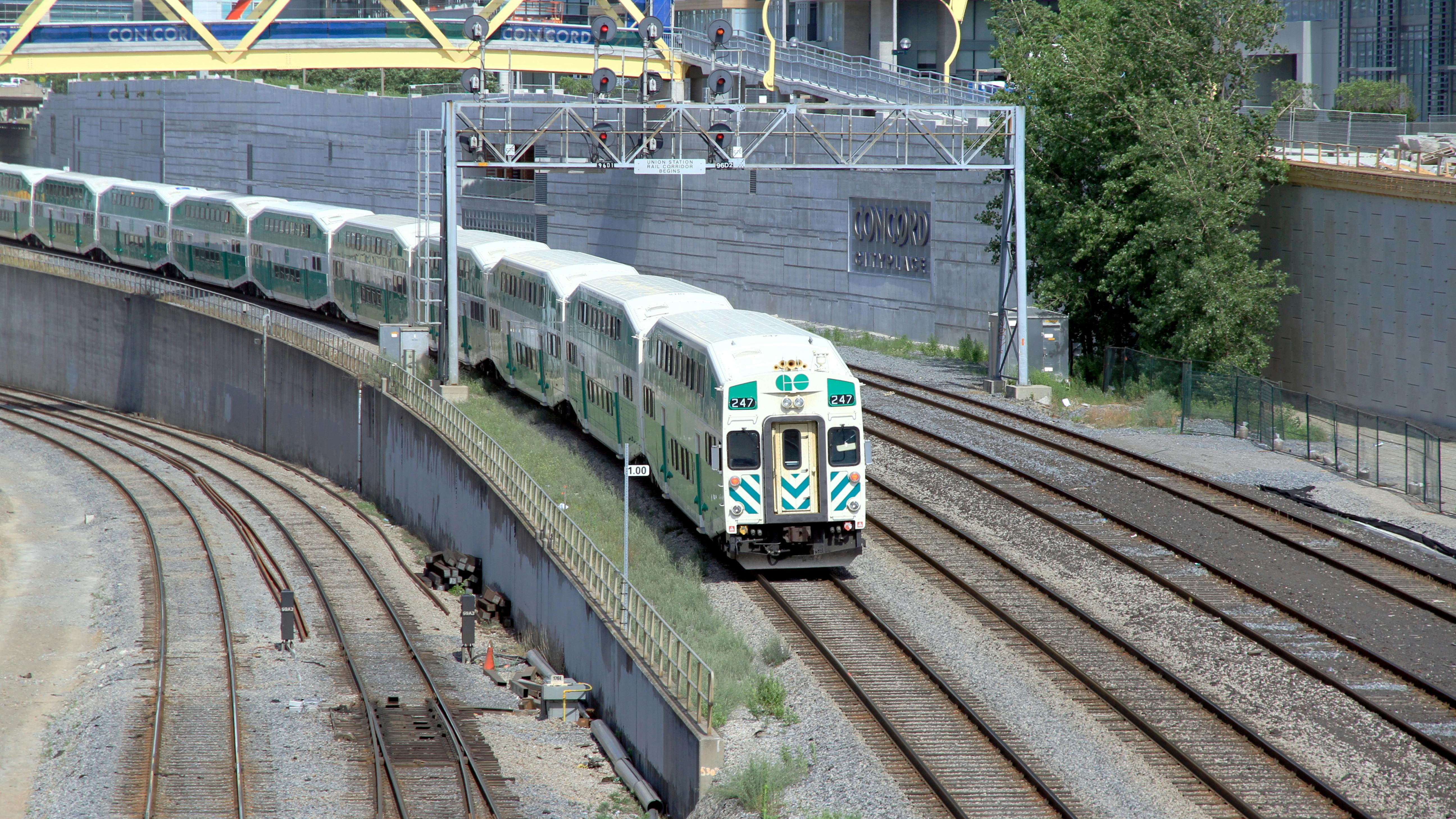 A GO Transit bi-level commuter rail train approaches on a curved, multi-track corridor in downtown Toronto, with the Concord CityPlace development visible in the background. A yellow signal gantry spans the tracks overhead, and several additional parallel tracks are visible in the foreground.