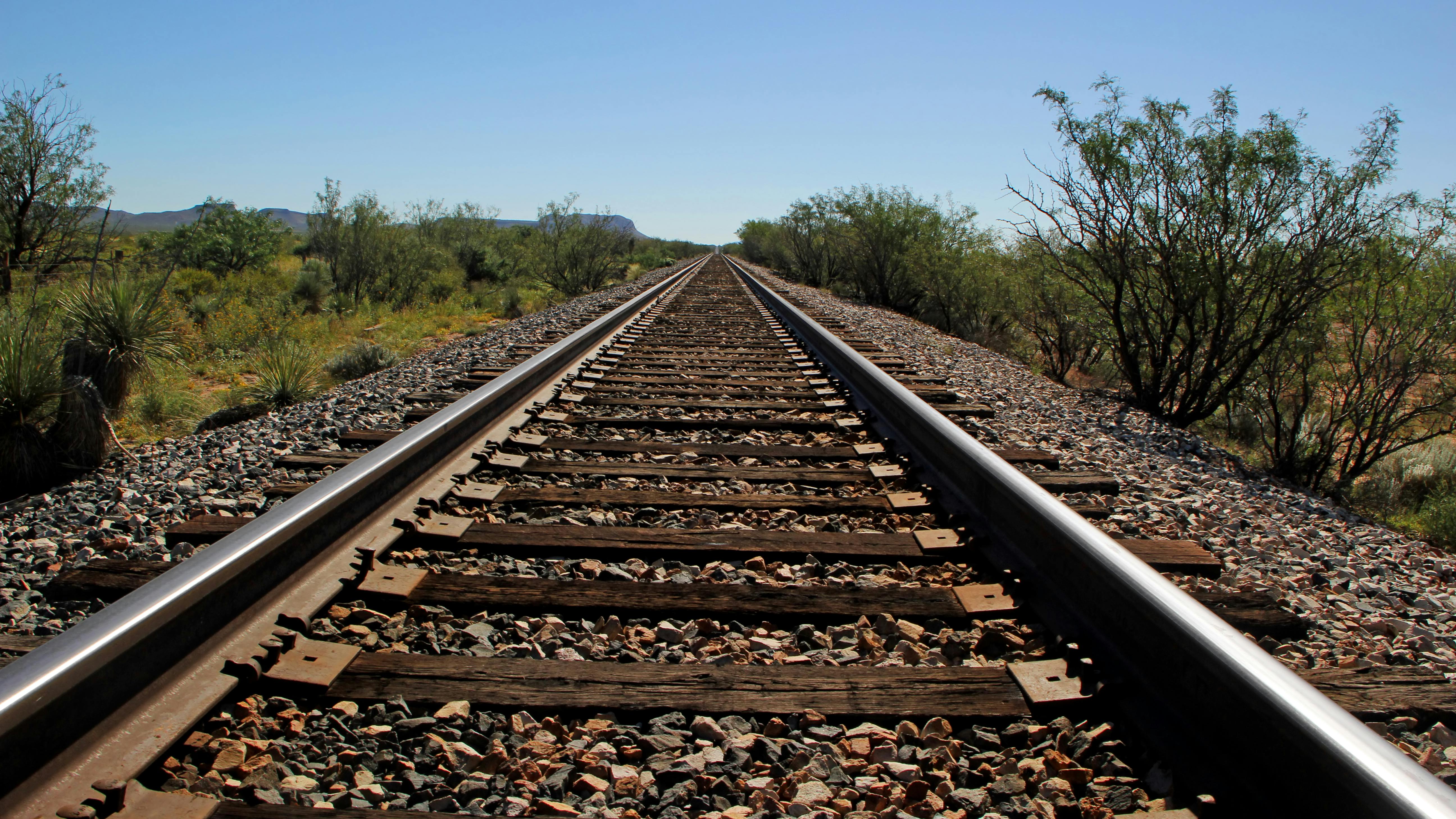 Railroad tracks extend straight toward the horizon through an arid desert landscape under a clear blue sky, with scrub brush, yucca plants and distant mountains flanking both sides of the track. Wooden ties and gravel ballast are visible in the foreground.