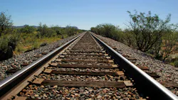Railroad tracks extend straight toward the horizon through an arid desert landscape under a clear blue sky, with scrub brush, yucca plants and distant mountains flanking both sides of the track. Wooden ties and gravel ballast are visible in the foreground. Railroad tracks extend straight toward the horizon through an arid desert landscape under a clear blue sky, with scrub brush, yucca plants and distant mountains flanking both sides of the track. Wooden ties and gravel ballast are visible in the foreground.