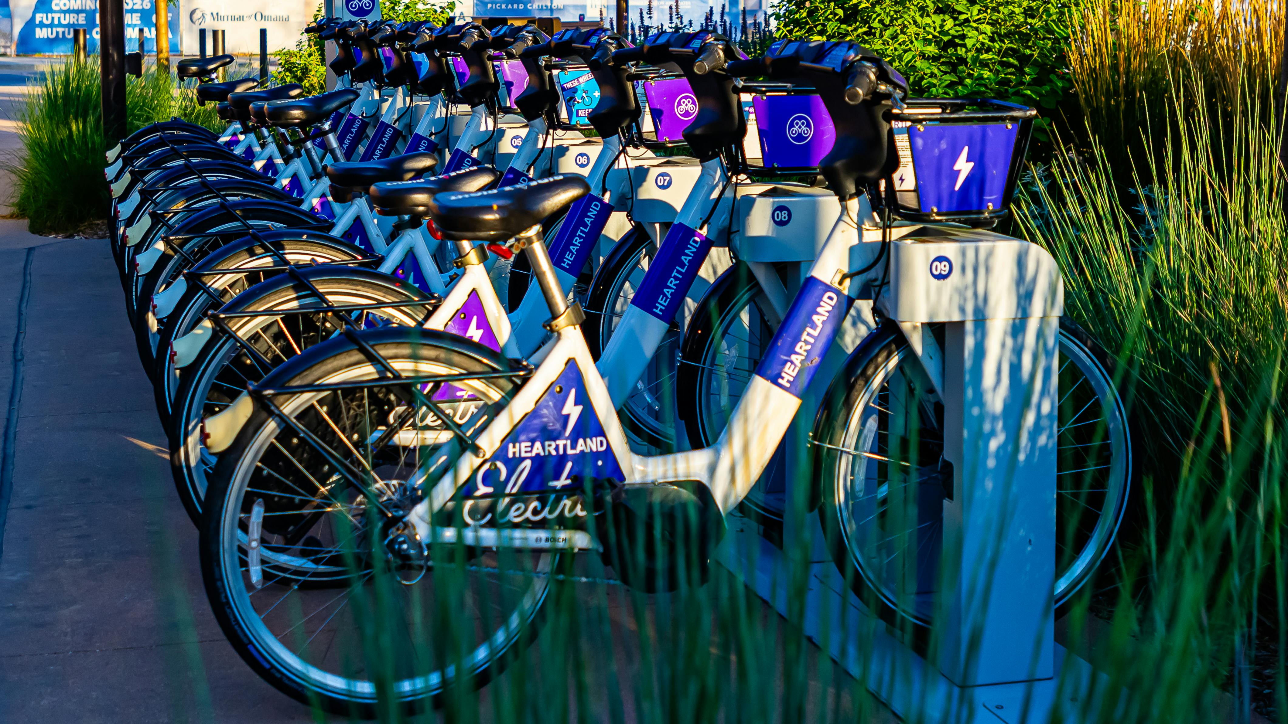 A row of Heartland B-cycle electric bike-share bicycles fills a docking station on a city sidewalk, their white frames and purple-and-blue branding prominently displayed. Each bike is numbered and equipped with a front basket. Ornamental grasses and trees frame the station, with urban construction signage visible in the background.