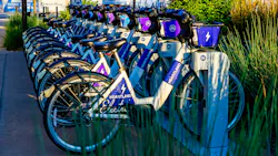 A row of Heartland B-cycle electric bike-share bicycles fills a docking station on a city sidewalk, their white frames and purple-and-blue branding prominently displayed. Each bike is numbered and equipped with a front basket. Ornamental grasses and trees frame the station, with urban construction signage visible in the background. A row of Heartland B-cycle electric bike-share bicycles fills a docking station on a city sidewalk, their white frames and purple-and-blue branding prominently displayed. Each bike is numbered and equipped with a front basket. Ornamental grasses and trees frame the station, with urban construction signage visible in the background.