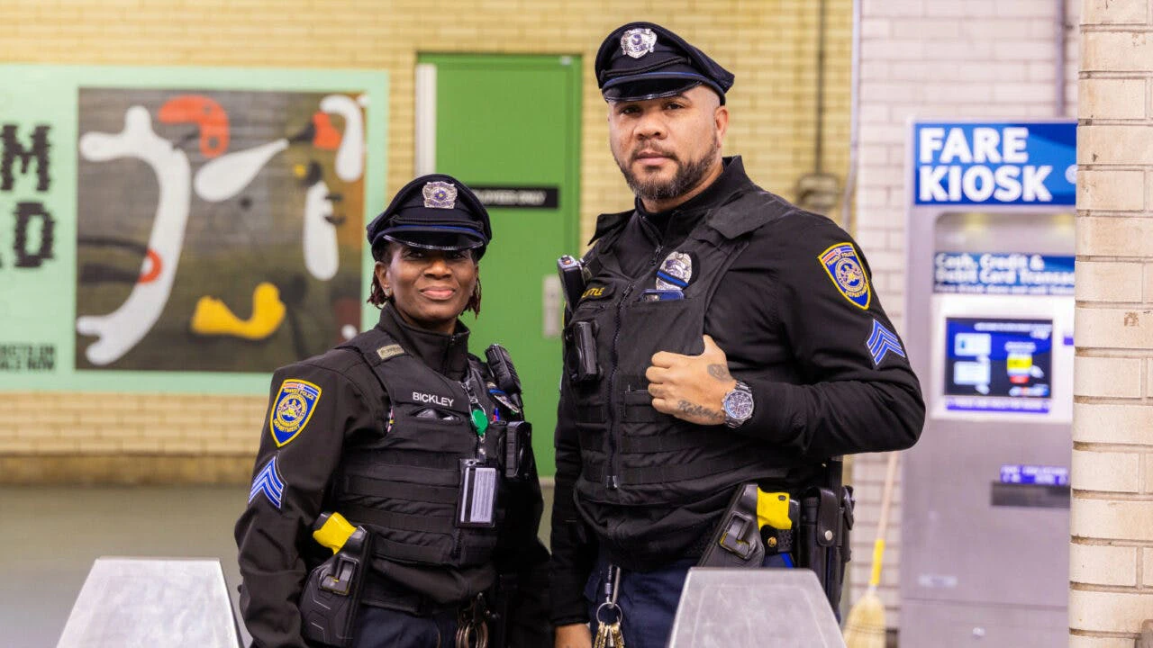 Two Southeastern Pennsylvania Transportation Authority (SEPTA) Transit Police officers stand near a fare kiosk inside a SEPTA station. Both officers wear black uniforms with body armor, duty belts, and SEPTA Transit Police Department patches. The officer on the left, identified by a nameplate as Bickley, smiles at the camera. The officer on the right, identified as Little, stands with one hand on his vest. A colorful mural and a green door are visible in the background.