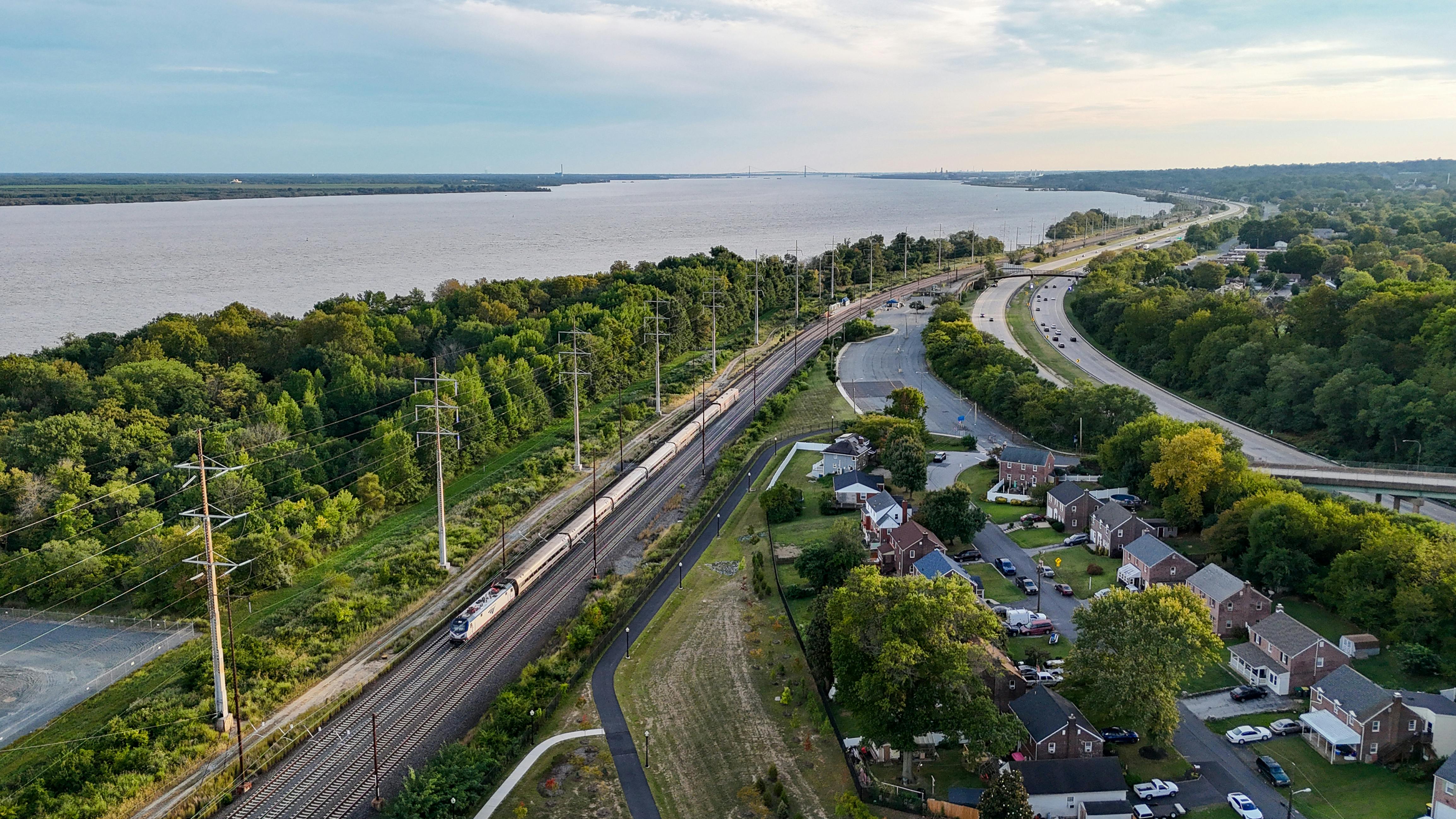 An aerial view of an Amtrak passenger train traveling along an electrified rail corridor beside a wide river. The double-track line runs through a tree-lined stretch flanked by a residential neighborhood of brick homes to the right and dense vegetation along the riverbank to the left. A curved roadway runs parallel to the tracks, and a bridge is visible in the distance where the river narrows toward the horizon. The scene is photographed during golden hour under a partly cloudy sky.