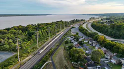 An aerial view of an Amtrak passenger train traveling along an electrified rail corridor beside a wide river. The double-track line runs through a tree-lined stretch flanked by a residential neighborhood of brick homes to the right and dense vegetation along the riverbank to the left. A curved roadway runs parallel to the tracks, and a bridge is visible in the distance where the river narrows toward the horizon. The scene is photographed during golden hour under a partly cloudy sky.