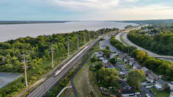 An aerial view of an Amtrak passenger train traveling along an electrified rail corridor beside a wide river. The double-track line runs through a tree-lined stretch flanked by a residential neighborhood of brick homes to the right and dense vegetation along the riverbank to the left. A curved roadway runs parallel to the tracks, and a bridge is visible in the distance where the river narrows toward the horizon. The scene is photographed during golden hour under a partly cloudy sky. An aerial view of an Amtrak passenger train traveling along an electrified rail corridor beside a wide river. The double-track line runs through a tree-lined stretch flanked by a residential neighborhood of brick homes to the right and dense vegetation along the riverbank to the left. A curved roadway runs parallel to the tracks, and a bridge is visible in the distance where the river narrows toward the horizon. The scene is photographed during golden hour under a partly cloudy sky.