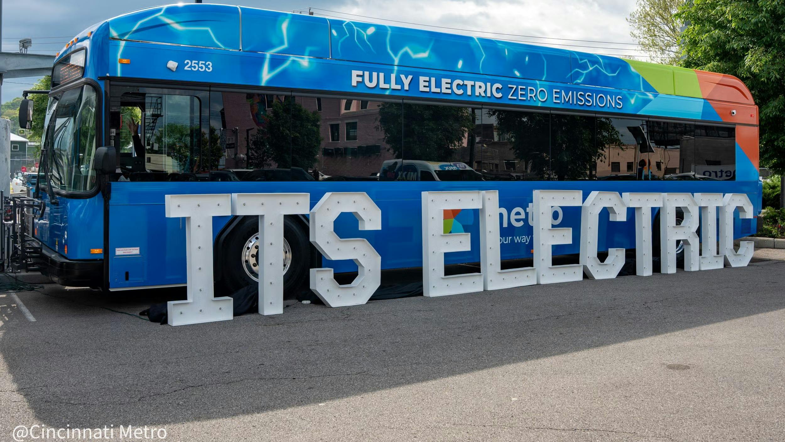 A Cincinnati Metro battery-electric bus (fleet number 2553) is parked outdoors at a launch event. The bus features a blue wrap with lightning bolt graphics and 'Fully Electric Zero Emissions' lettering along the upper side, along with a multicolored stripe &mdash; blue, green, and orange &mdash; along the rear. Large white marquee-style letters spelling 'ITS ELECTRIC' are arranged in front of the bus, partially obscuring the Metro logo on the vehicle's side. The Cincinnati Metro logo is visible in the lower-left corner of the image.