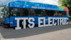 A Cincinnati Metro battery-electric bus (fleet number 2553) is parked outdoors at a launch event. The bus features a blue wrap with lightning bolt graphics and 'Fully Electric Zero Emissions' lettering along the upper side, along with a multicolored stripe — blue, green, and orange — along the rear. Large white marquee-style letters spelling 'ITS ELECTRIC' are arranged in front of the bus, partially obscuring the Metro logo on the vehicle's side. The Cincinnati Metro logo is visible in the lower-left corner of the image. A Cincinnati Metro battery-electric bus (fleet number 2553) is parked outdoors at a launch event. The bus features a blue wrap with lightning bolt graphics and 'Fully Electric Zero Emissions' lettering along the upper side, along with a multicolored stripe — blue, green, and orange — along the rear. Large white marquee-style letters spelling 'ITS ELECTRIC' are arranged in front of the bus, partially obscuring the Metro logo on the vehicle's side. The Cincinnati Metro logo is visible in the lower-left corner of the image.