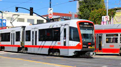 A San Francisco Municipal Transportation Agency (SFMTA) Muni light-rail vehicle, car number 2004B, travels through a street-level intersection in San Francisco on a sunny day. The train's silver and red livery with the Muni wordmark is visible along the side. A second Muni train is partially visible to the right. Traffic signals, street signs — including a 'Do Not Enter' sign — and commercial buildings line the background under a clear blue sky.