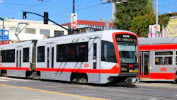 A San Francisco Municipal Transportation Agency (SFMTA) Muni light-rail vehicle, car number 2004B, travels through a street-level intersection in San Francisco on a sunny day. The train's silver and red livery with the Muni wordmark is visible along the side. A second Muni train is partially visible to the right. Traffic signals, street signs — including a 'Do Not Enter' sign — and commercial buildings line the background under a clear blue sky. A San Francisco Municipal Transportation Agency (SFMTA) Muni light-rail vehicle, car number 2004B, travels through a street-level intersection in San Francisco on a sunny day. The train's silver and red livery with the Muni wordmark is visible along the side. A second Muni train is partially visible to the right. Traffic signals, street signs — including a 'Do Not Enter' sign — and commercial buildings line the background under a clear blue sky.