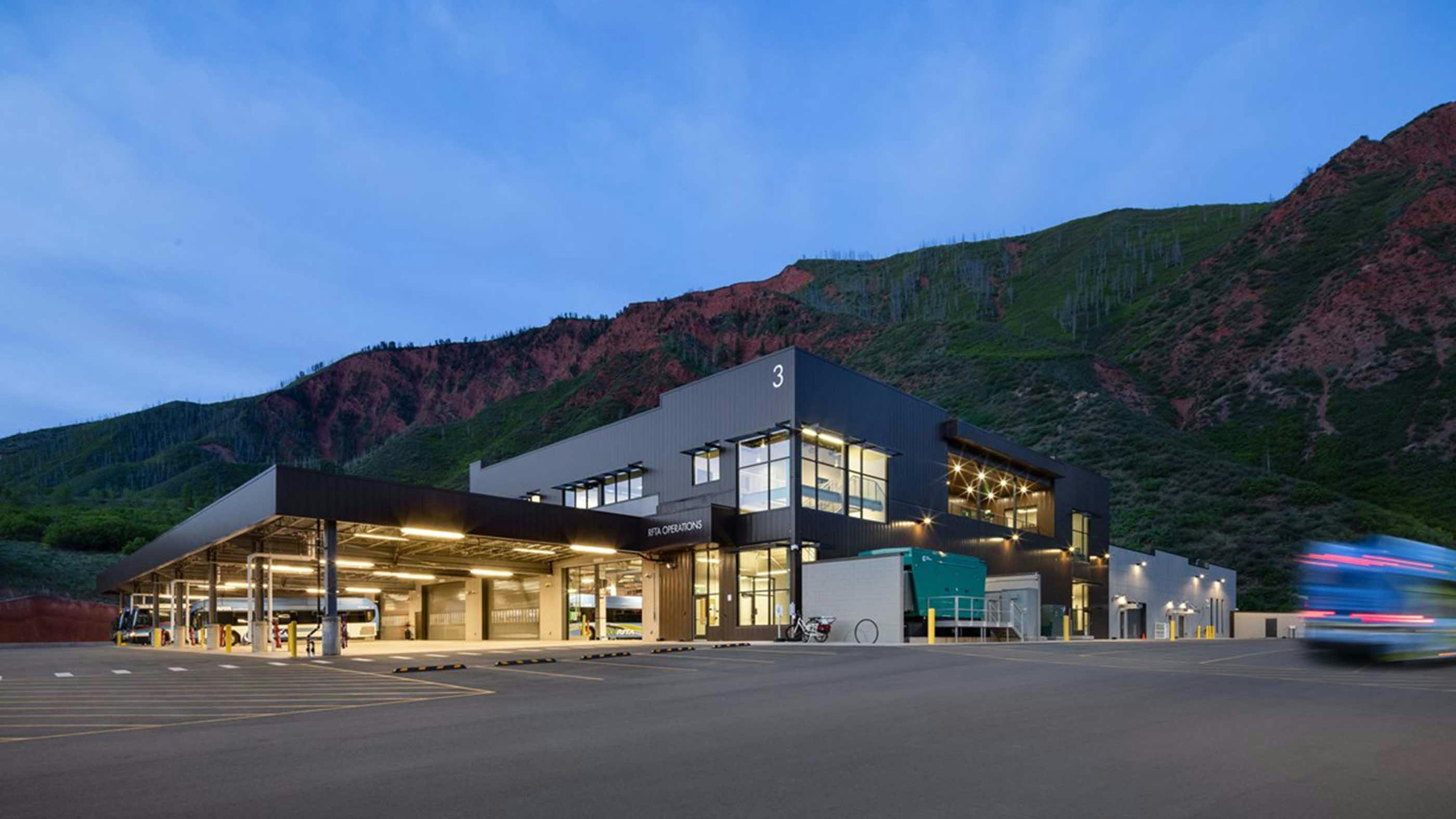 The exterior of the Roaring Fork Transportation Authority (RFTA) Operations facility &mdash; a modern, multi-story maintenance and operations building with dark metal panel cladding and large illuminated windows &mdash; is photographed at dusk. An open bus maintenance bay stretches along the left side of the building, with several buses visible inside under bright overhead lighting. A bus passes in motion blur at the right edge of the frame. The facility is set against a dramatic backdrop of red rock and green hillside terrain under a deep blue twilight sky.