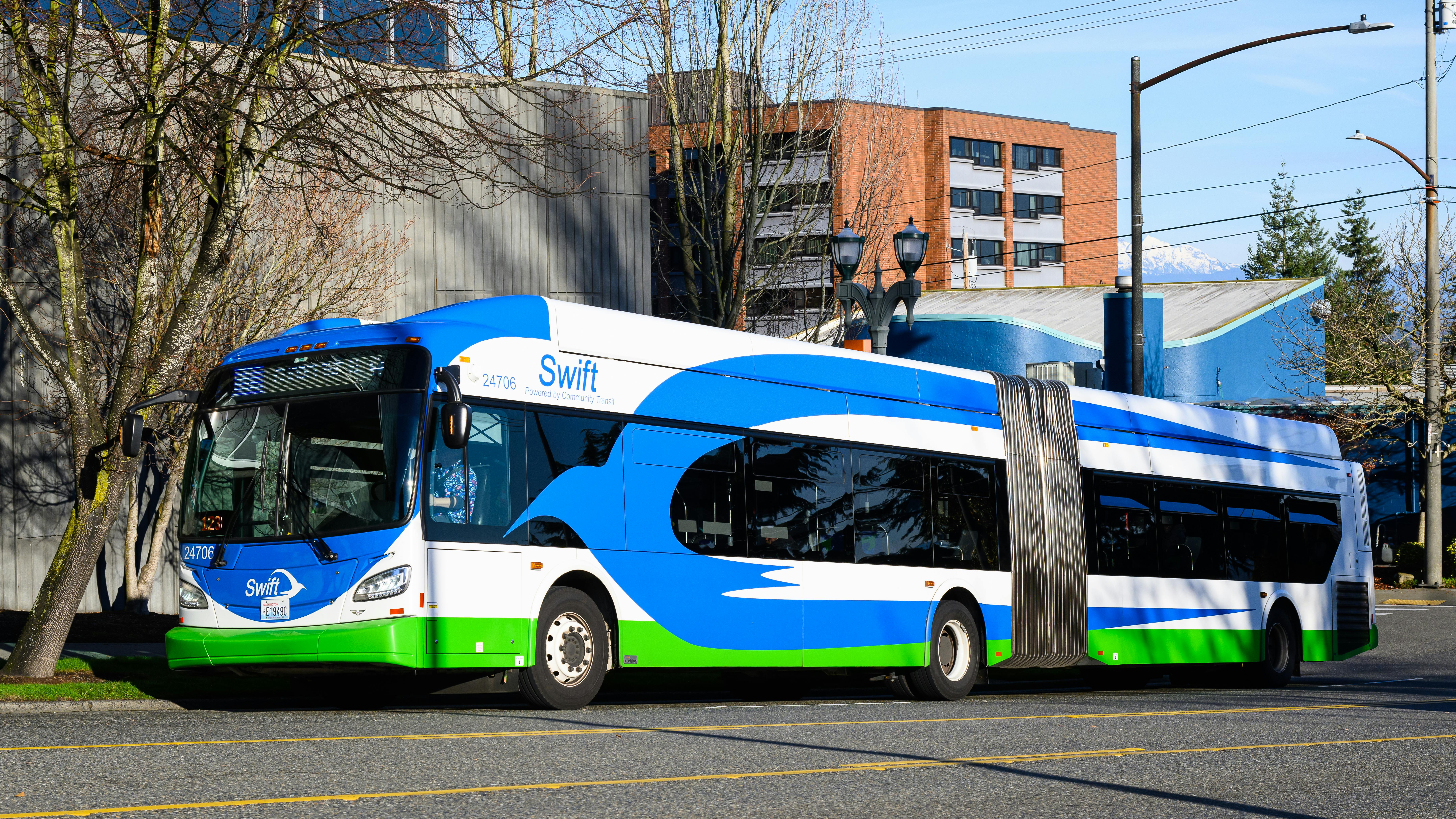 A Community Transit Swift bus rapid transit articulated bus, numbered 24706, travels along a city street in the Pacific Northwest. The bus features the Swift BRT livery of blue, white and green with a wave design. Snow-capped mountains are visible in the background under a clear blue sky.