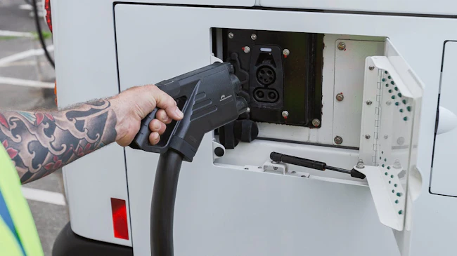 A transit worker in a high-visibility safety vest uses both hands to connect a large DC fast-charging connector to the rear charge port of a white battery-electric bus. The worker's arm is visibly tattooed. The charge port door is open, revealing the vehicle's charging inlet.