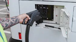 A transit worker in a high-visibility safety vest uses both hands to connect a large DC fast-charging connector to the rear charge port of a white battery-electric bus. The worker's arm is visibly tattooed. The charge port door is open, revealing the vehicle's charging inlet.