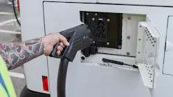 A transit worker in a high-visibility safety vest uses both hands to connect a large DC fast-charging connector to the rear charge port of a white battery-electric bus. The worker's arm is visibly tattooed. The charge port door is open, revealing the vehicle's charging inlet. A transit worker in a high-visibility safety vest uses both hands to connect a large DC fast-charging connector to the rear charge port of a white battery-electric bus. The worker's arm is visibly tattooed. The charge port door is open, revealing the vehicle's charging inlet.