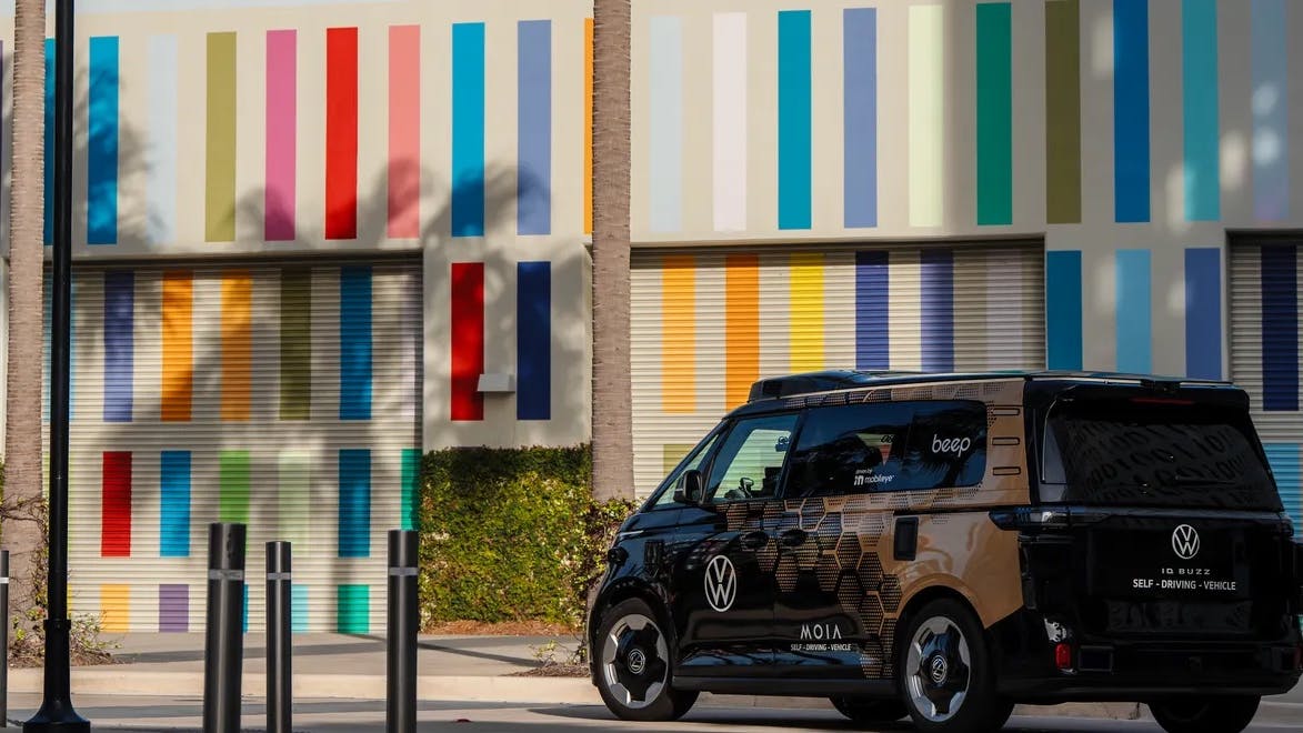 A Volkswagen ID. Buzz self-driving vehicle operated by MOIA and powered by Mobileye autonomy technology, branded with 'beep' signage, is parked near a building with a colorful multicolored striped exterior mural. The vehicle features a dark honeycomb-patterned wrap and roof-mounted sensor equipment.