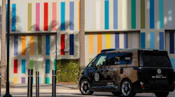 A Volkswagen ID. Buzz self-driving vehicle operated by MOIA and powered by Mobileye autonomy technology, branded with 'beep' signage, is parked near a building with a colorful multicolored striped exterior mural. The vehicle features a dark honeycomb-patterned wrap and roof-mounted sensor equipment.