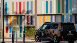 A Volkswagen ID. Buzz self-driving vehicle operated by MOIA and powered by Mobileye autonomy technology, branded with 'beep' signage, is parked near a building with a colorful multicolored striped exterior mural. The vehicle features a dark honeycomb-patterned wrap and roof-mounted sensor equipment. A Volkswagen ID. Buzz self-driving vehicle operated by MOIA and powered by Mobileye autonomy technology, branded with 'beep' signage, is parked near a building with a colorful multicolored striped exterior mural. The vehicle features a dark honeycomb-patterned wrap and roof-mounted sensor equipment.