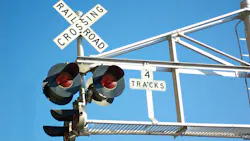 A railroad crossing signal assembly photographed from below against a clear blue sky. The X-shaped crossbuck sign reads 'Railroad Crossing' and a rectangular placard below reads '4 Tracks.' Three red warning lights are mounted on the signal mast, and a white metal overhead gantry extends to the right. A railroad crossing signal assembly photographed from below against a clear blue sky. The X-shaped crossbuck sign reads 'Railroad Crossing' and a rectangular placard below reads '4 Tracks.' Three red warning lights are mounted on the signal mast, and a white metal overhead gantry extends to the right.