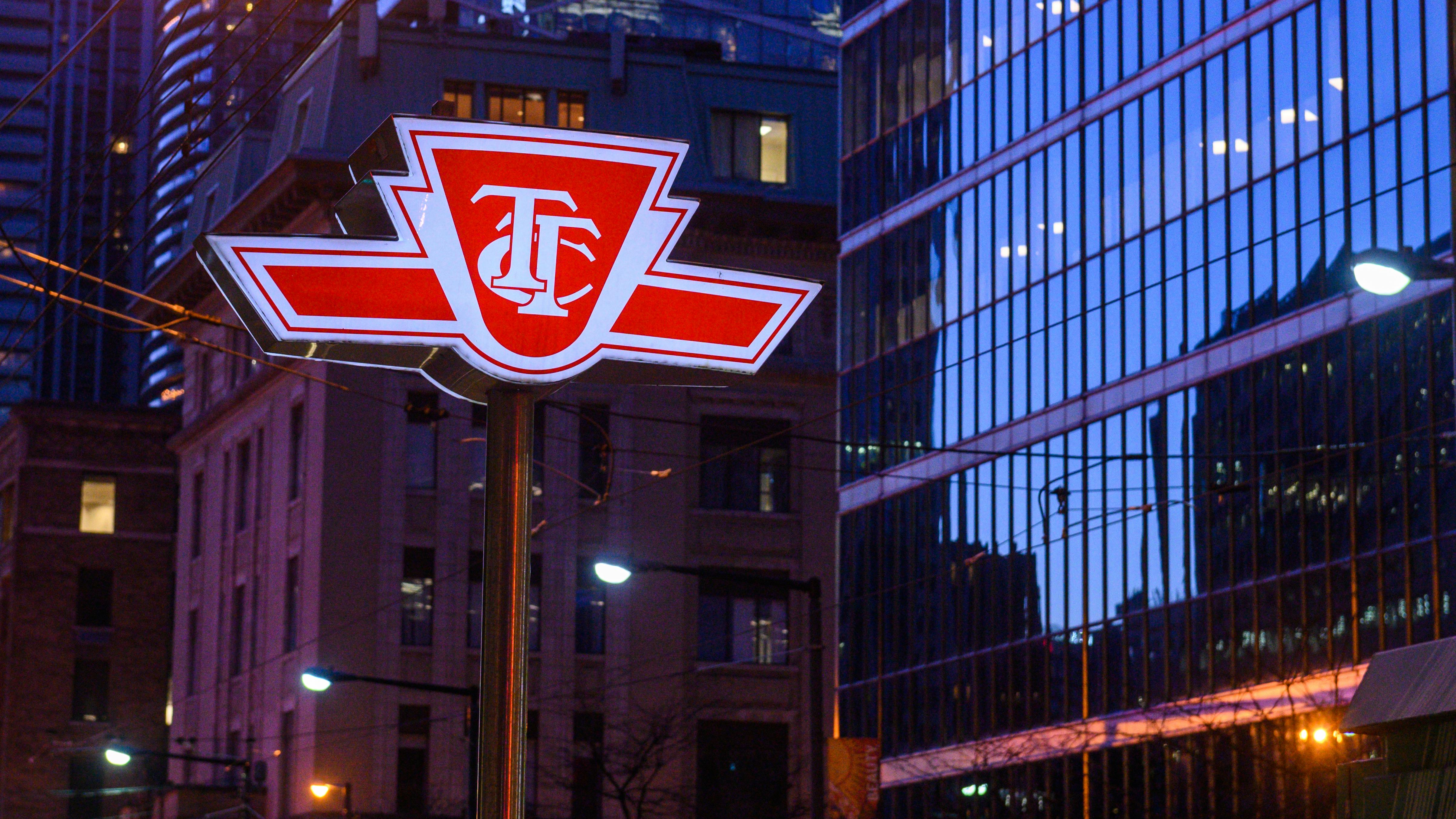 The illuminated red-and-white logo sign of the Toronto Transit Commission (TTC) mounted on a pole at street level, photographed at dusk or night in a downtown urban setting. Glass-clad high-rise buildings and a historic brick building are visible in the background, with streetlights and overhead wires reflecting in the glass facade.