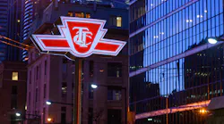 The illuminated red-and-white logo sign of the Toronto Transit Commission (TTC) mounted on a pole at street level, photographed at dusk or night in a downtown urban setting. Glass-clad high-rise buildings and a historic brick building are visible in the background, with streetlights and overhead wires reflecting in the glass facade.
