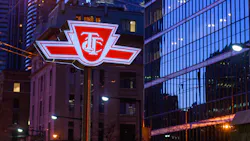 The illuminated red-and-white logo sign of the Toronto Transit Commission (TTC) mounted on a pole at street level, photographed at dusk or night in a downtown urban setting. Glass-clad high-rise buildings and a historic brick building are visible in the background, with streetlights and overhead wires reflecting in the glass facade. The illuminated red-and-white logo sign of the Toronto Transit Commission (TTC) mounted on a pole at street level, photographed at dusk or night in a downtown urban setting. Glass-clad high-rise buildings and a historic brick building are visible in the background, with streetlights and overhead wires reflecting in the glass facade.