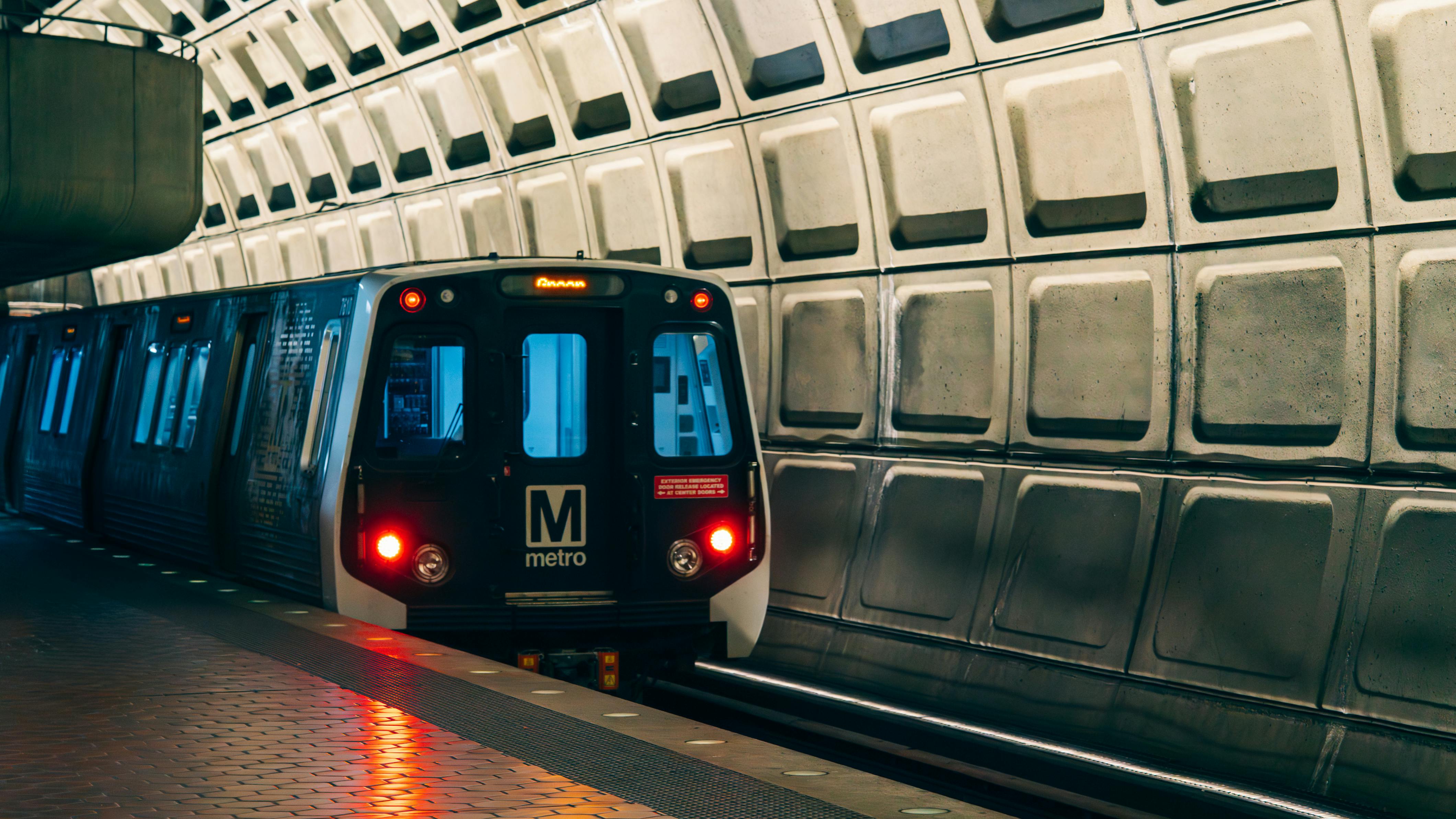 A WMATA Metrorail car rests at a station platform, facing the camera with its red headlights on and a destination displayed on the LED header board. The car bears the agency's block 'M metro' logo on its nose. The platform floor features tactile edge strips, and the station's iconic coffered concrete barrel vault ceiling fills the upper right of the frame. The lighting gives the scene a cool blue-and-amber tone.