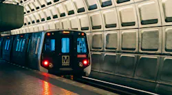 A WMATA Metrorail car rests at a station platform, facing the camera with its red headlights on and a destination displayed on the LED header board. The car bears the agency's block 'M metro' logo on its nose. The platform floor features tactile edge strips, and the station's iconic coffered concrete barrel vault ceiling fills the upper right of the frame. The lighting gives the scene a cool blue-and-amber tone.