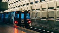 A WMATA Metrorail car rests at a station platform, facing the camera with its red headlights on and a destination displayed on the LED header board. The car bears the agency's block 'M metro' logo on its nose. The platform floor features tactile edge strips, and the station's iconic coffered concrete barrel vault ceiling fills the upper right of the frame. The lighting gives the scene a cool blue-and-amber tone. A WMATA Metrorail car rests at a station platform, facing the camera with its red headlights on and a destination displayed on the LED header board. The car bears the agency's block 'M metro' logo on its nose. The platform floor features tactile edge strips, and the station's iconic coffered concrete barrel vault ceiling fills the upper right of the frame. The lighting gives the scene a cool blue-and-amber tone.